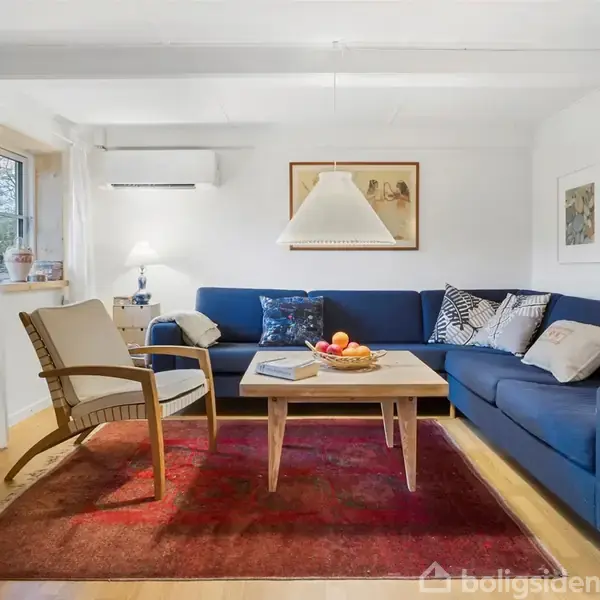 En stue med en blå hjørnesofa i fokus. En træstol og sofabord står på et rødt tæppe. En lampe hænger over sofabordet. Værelset har hvide vægge og trægulv.