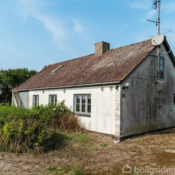 Et ældre, hvidt hus med skråt tag og antenne står på en bar, jordet grund omkranset af buske og træer under en blå himmel.