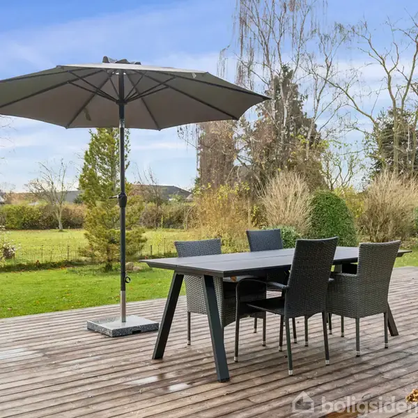 Et bord med stole står på en træterrasse; en stor parasol giver skygge. Baggrunden består af en frodig have med græsplæne og buske under en blå himmel.