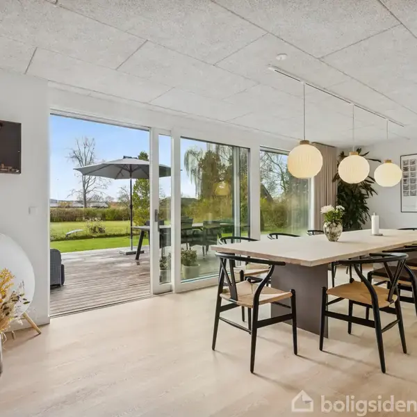En spisestue med et stort spisebord omgivet af otte stole. Bordet er dekoreret med moderne lamper. Rummet har glasdøre, der åbner ud til en terrasse og en have.