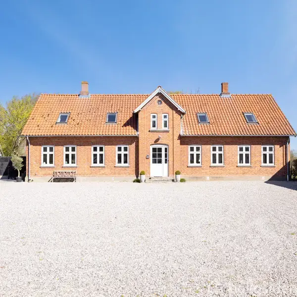 En rødstensbygning med rødt tegltag står i en grusbelagt gårdsplads. Bygningen har flere vinduer, og der er et mindre træhus i baggrunden. Solskin kaster lys over scenen.