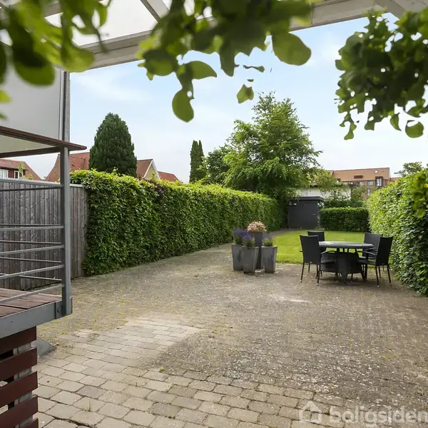 Terrasse med stenbelægning, et bord og seks stole står i centrum. Omgivet af hække og forhaver. Grønne planter hænger ned fra loftet, hvilket skaber en hyggelig atmosfære.