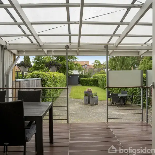 Overdækket terrasse med spisebord og stole. Terrassen åbner sig mod en velplejet have med grussti, græsplæne og omkransende buske.