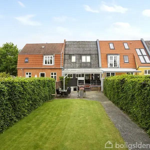 Rækkehus med en orange og grå facade står i midten, omkranset af velplejede hække og en græsplæne foran. Der er havemøbler på terrassen, og himlen er blå.
