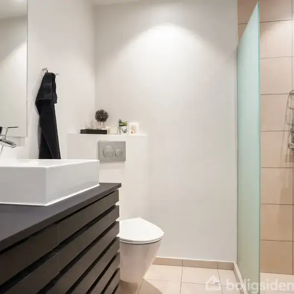 Håndvask med sort bordplade står ved siden af et toilet. En brusekabine er i baggrunden. Rummet er lyst og moderne med hvide vægge og fliser.