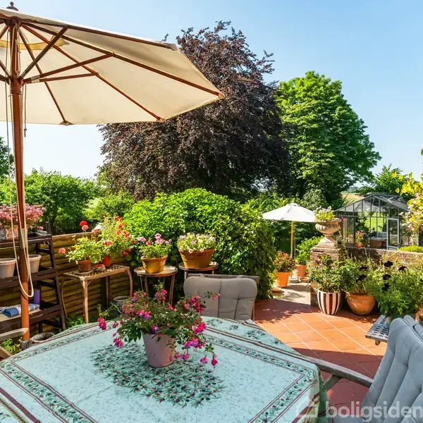 En terrasse med et bord dækket af en dug og blomster, omkranset af stole. I baggrunden er der planter, store træer og en parasol, skaber en hyggelig haveatmosfære.