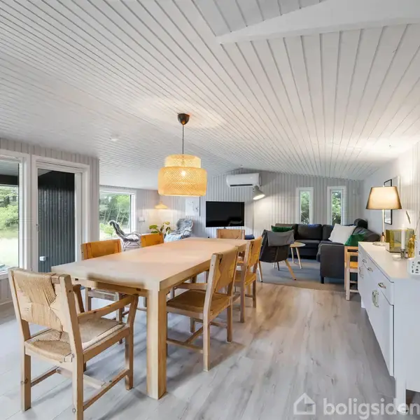 Spisebord med træstole står i en lys stue med hvide trævægge og -loft. En lampe hænger over bordet. Stuen har store vinduer, moderne møbler og udsigt til en have.