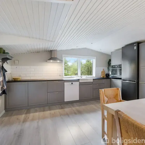 Et moderne køkken med grå skabe og hvide vægge. Der er en opvaskemaskine og et stort køleskab. Et spisebord med stole står i forgrunden.