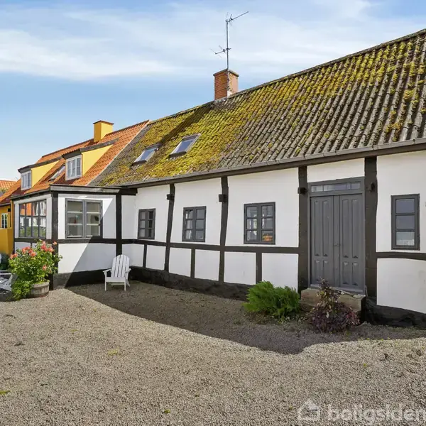 Traditionelt bindingsværkshus med hvidmalet facade og mosbevokset tegltag ses stille stående. På gårdspladsen er der grus, en hvid stol og planter. Enkelt nummer "3" på væggen indikerer adressen.