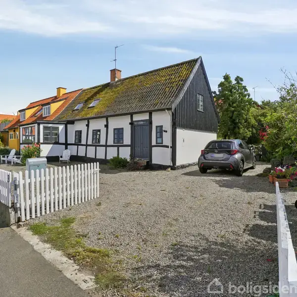 Et bindingsværkshus med sort træværk, bil parkeret foran på grus, hvidt stakit omkring terrasse med havemøbler, placeret i en landsby med flere farverige huse langs en vej.