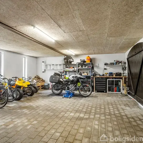 Cykler og motorcykler parkeret på et flisebelagt gulv, omgivet af værktøj og hylder i en godt oplyst garage med åbne trædøre.