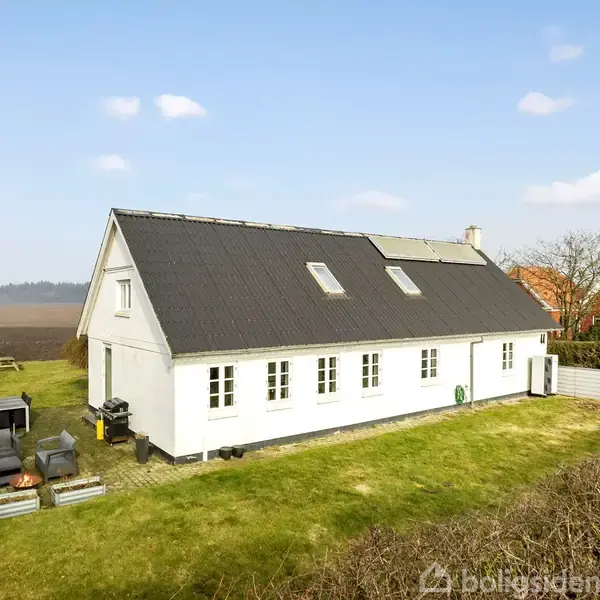 Hvidt hus med mørkt tag står på græsplæne i åbent landskab med et par træer i nærheden og marker i baggrunden. Haveredskaber og møbler er synlige til venstre.