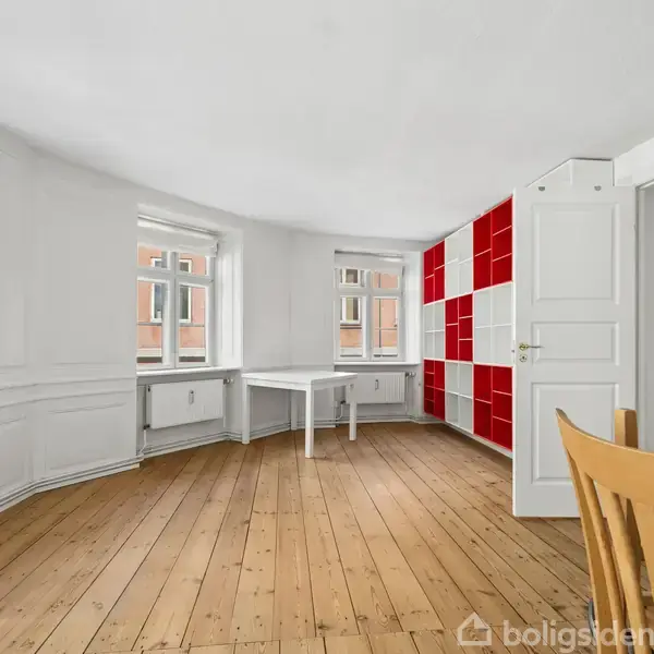 Et lyst rum med trægulv, en hvid rejol med røde firkantede paneler. Et bord står ved to store vinduer, der lukker lys ind.