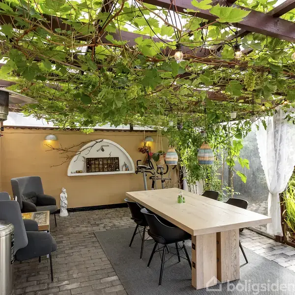 Under et pergola med grønne ranker står et langt, lyst træbord omgivet af stole. To farverige lamper hænger over bordet, og en terrassevarmer står i hjørnet.