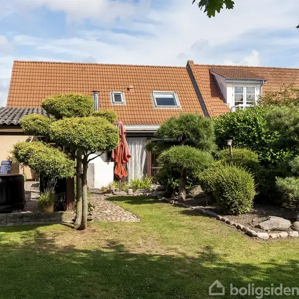 En have med veltrimmede buske og træer foran et hus med rødt tegltag og flere ovenlysvinduer. En parasol står ved en terrasse med stenbelægning under en blå himmel.