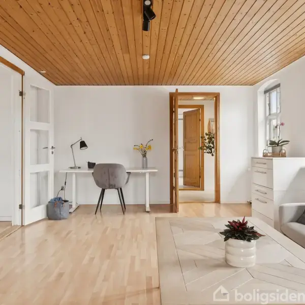 Stue med lyst trægulv og træloft. Skrivebord med stol står op ad en hvid væg. Sofa og plante i venstre side. Store vinduer lyser rummet op.