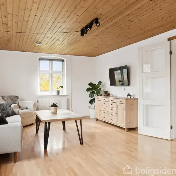 En lys stue med hvidmalede vægge og lyse trægulve. En sofa og sofabord dominerer rummet, flankeret af en skænk og en plante. Et vindue lader naturligt lys ind.