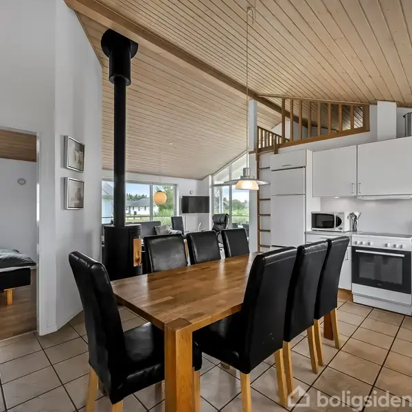 Spisebord med seks sorte stole står i en moderne, lys stue med åben køkkenindretning, hvide skabe og hvidevarer. Træloft og gulvfliser skaber en hyggelig atmosfære. En seng ses i et tilstødende værelse.