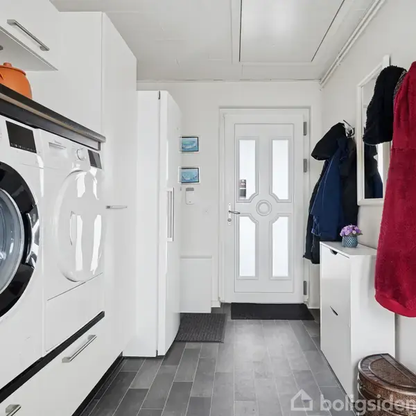 Vaskemaskine og tørretumbler står i en bryggersindgang med gråt flisegulv. På væggen hænger frakker, og døren fører ud til det fri. Tre små hylder ses over et hvidt skab.