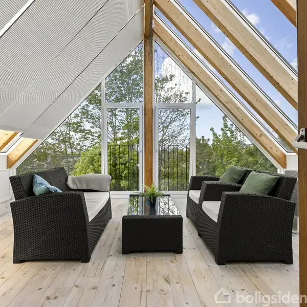 To sorte kurvemøbler, placeret på trægulv, vender mod hinanden med et lille bord imellem. Store skråvinduer giver udsigt til grønne træer og en klar himmel.