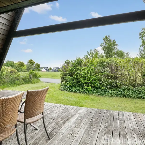 Terrassemøbler står på en træterrasse, muligvis en del af et sommerhus. De er placeret ud mod en grøn have med buske og græs, og i baggrunden ses huse.