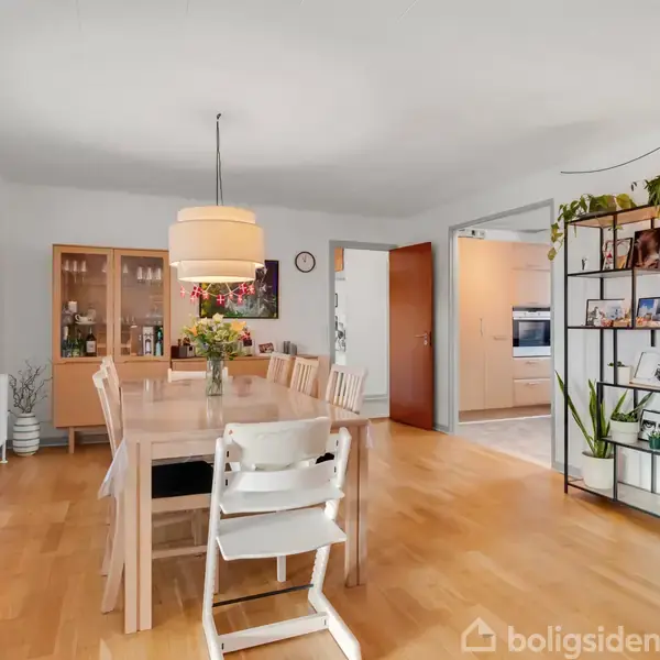 Et spisebord med stole står i centrum af et lyst rum. Væggene har hylder og planter, og store vinduer slipper lys ind. I baggrunden ses en reol og dør til køkken.