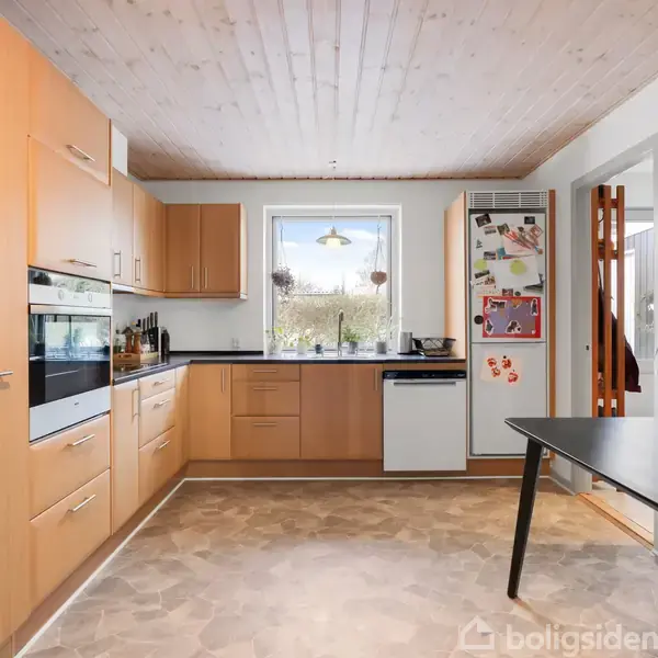 Køkken med træskabe står mod en væg, opvaskemaskine og ovn installeret. Bordet til højre ved siden af stole. Vindue med udsigt til have. Køleskab med magneter.