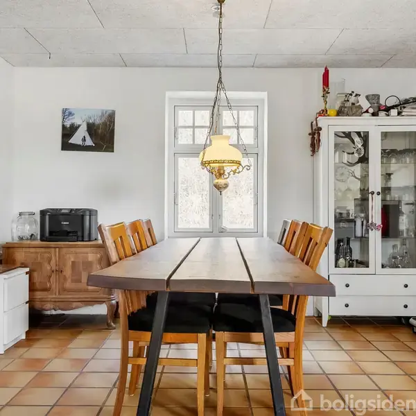 Et spisebord med seks stole står i et lyst rum. Der hænger en lampe over bordet. Et skab og et maleri pryder væggene, med vindue i baggrunden.