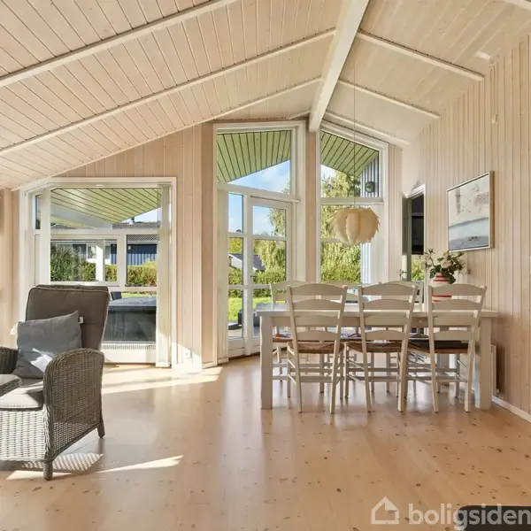 Stue med spisebord og stole, hvor flere sidder samlet. Rummet har trævæggene, højloftet trætag og store vinduer med udsigt til en have i sollys. En brændeovn står til højre.