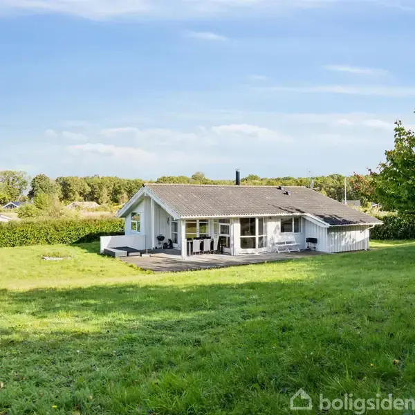 En hvidmalet bungalow med en veranda står i en velplejet have. Huset er omgivet af grønne træer og en hæk med en blå himmel i baggrunden.