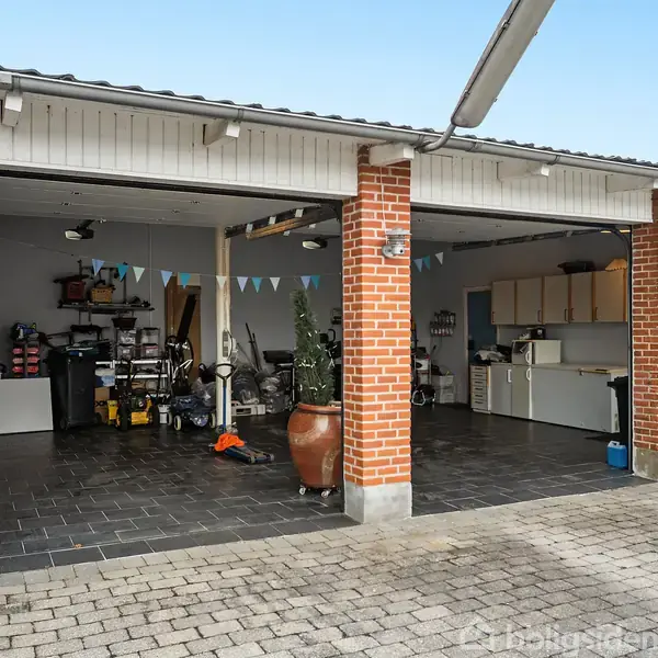 Garage med to åbne porte. Indenfor står værktøj og udstyr organiseret langs væggen; et lille køkkenområde ses i det ene hjørne. Farvede guirlander hænger fra loftet.