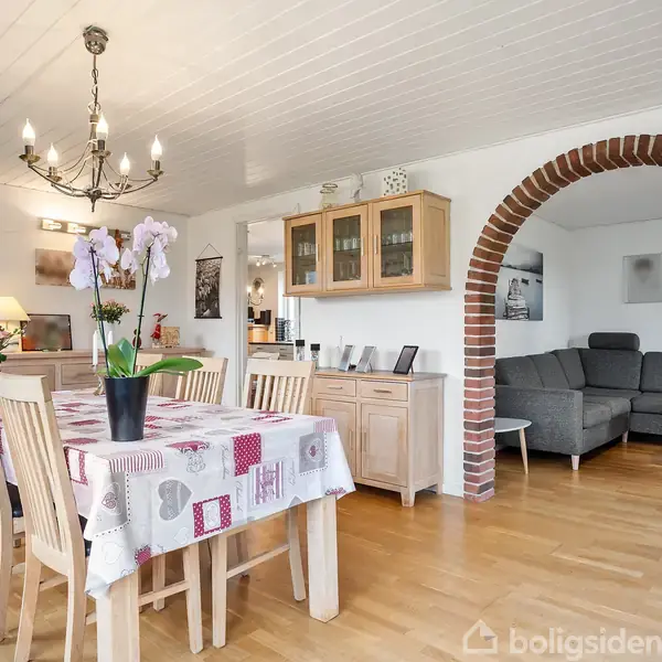 Spisebord dækket med dug og blomster, omgivet af træstole, står i en spisestue. En lysestage hænger over bordet. I baggrunden ses en grå sofa og en bueformet murstensåbning.