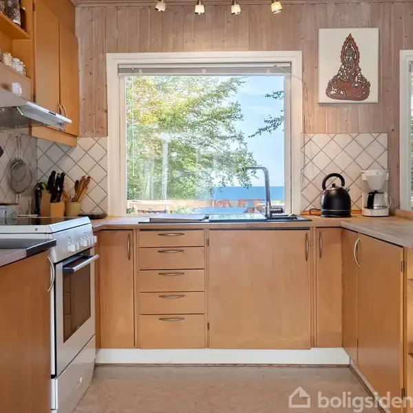 Køkken med lyse træskabe omkring en vask. Udsigt til havet gennem et vindue. Kogegrej og brød på hylder. Flisebagvæg og maleri. Bordplade med kedel og kaffemaskine.