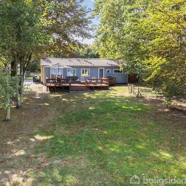 Huset står stille i en solrig have omgivet af tætte træer. Foran huset er en stor træterrasse med havemøbler. Græsplænen er lys og rummelig.