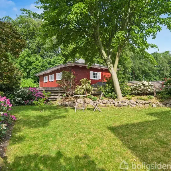 Rødt hus med hvide vinduer står i en frodig have. Blomster og buske omkranser græsplænen, mens et højt træ rejser sig opad. Overskyet himmel i baggrunden.
