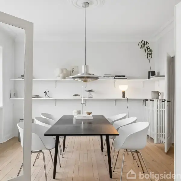 Et spisebord med seks hvide stole står i et lyst rum. Bordet er sort, og en lampe hænger over det. Hylder på bagvæggen indeholder bøger og dekoration.