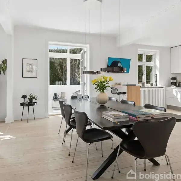 Spisebord omgivet af moderne stole står i lys og rummelig spisestue med trægulv. Bordet er dekoreret med planter og bøger. Stort vindue og hvidt køkken skaber en lys atmosfære.