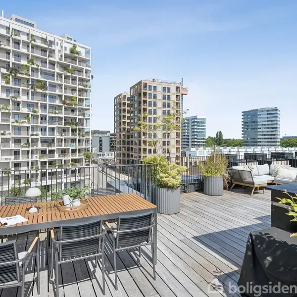 Tagterrasse med træbord og stole omringet af krukkeplanter. Moderne lejlighedsbygninger med altaner i baggrunden under en blå himmel.