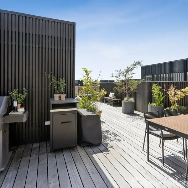 Grill og plantekrukker står på en moderne træterrasse. Der er udendørs spisebord med stole. Omgivet af sorte panelvægge og flere grønne planter, under en klar blå himmel.