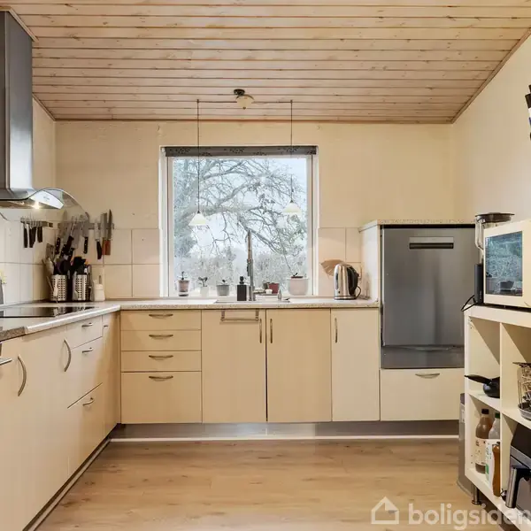 Køkken i lyst træ, med ovn og komfur på venstre side, opvaskemaskine på højre, og vindue med udsigt til træer i midten. Beige skabe og trægulv.