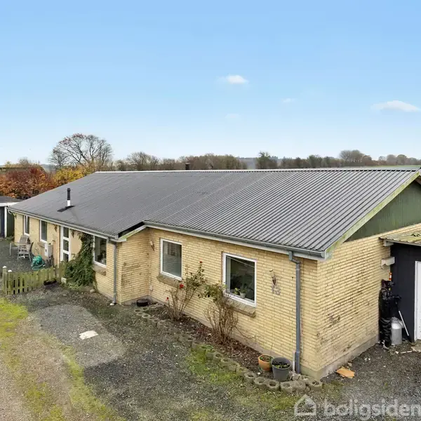 En gul murstensbygning med bølgetaget tag står stille på landet, omgivet af en grusvej og åbne marker.