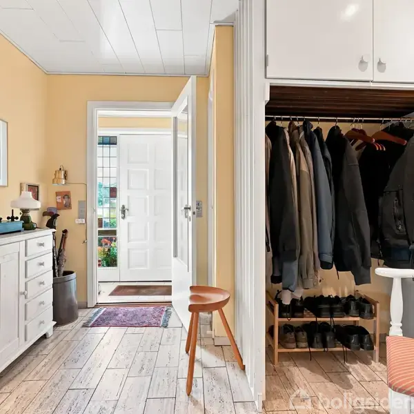 Garderobe med tøj på bøjler og sko nedenunder, står åben i en gang. På venstre side er der en hvid skænk med dekorationer. Yderligere ses en åben dør til huset.