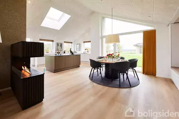 Spisebord med sorte stole står på et rundt tæppe i moderne stue med lyst trægulv. Store vinduer til venstre giver udsigt til grøn have. Lavt, sort pejs til venstre.