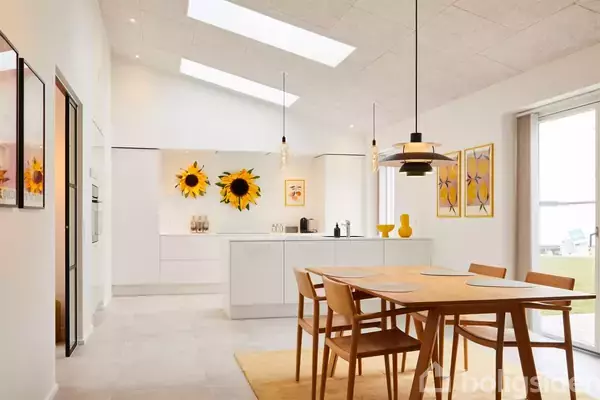 Et køkken-alrum har et lyst bord med fire stole i forgrunden. To store solsikkemalerier hænger over en hvid køkkenø i det soloplyste rum med moderne udsmykning.