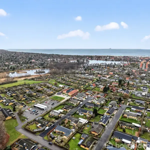Boligområde set fra luften, med huse spredt langs veje. Flere grønne områder omgiver bygningerne. I baggrunden ses en sø og en horisontlinje med hav og skov.