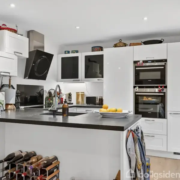 Et moderne køkken med hvide skabe og en sort bordplade, hvorpå der står en flaske og en skål med citroner. Der er indbyggede ovne og en vinkøler i baggrunden.