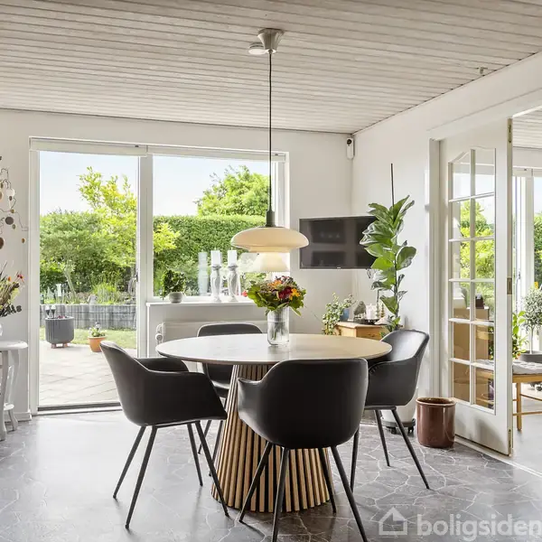 Rundt spisebord med fire sorte stole, blomster i midten; omgivet af store vinduer, lyse vægge, og en åben gårdhave i baggrunden med grønne planter udenfor.