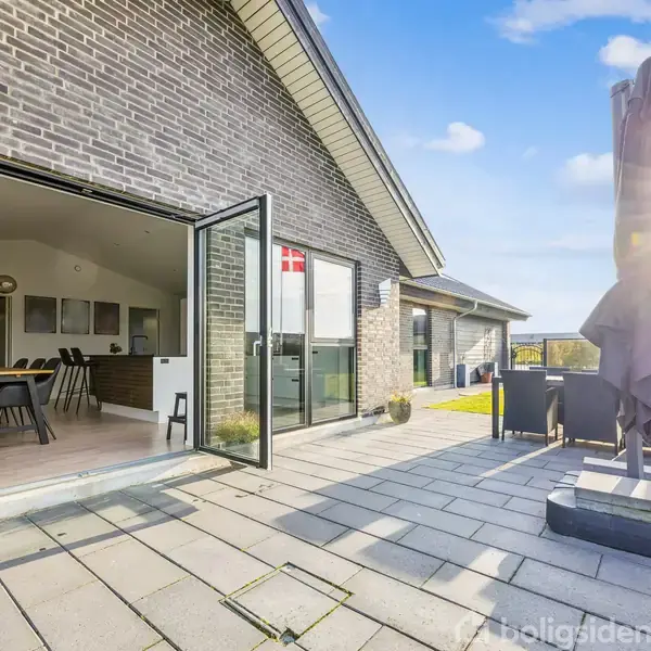 En moderne terrasse med åben skydedør ind til en spisestue, stol og bord på terrassen. En sammenfoldet parasol står til højre, omgivet af en græsplæne med klart blå himmel.