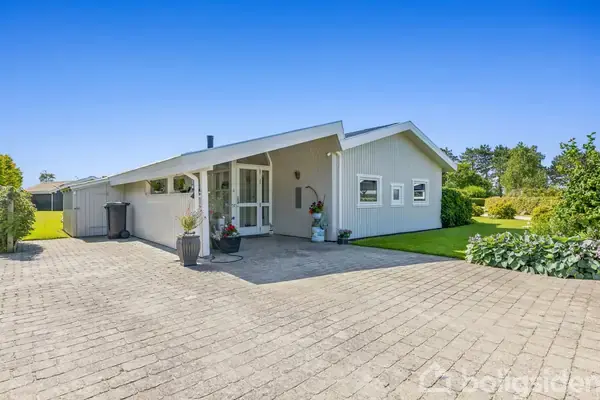 Et gråmalet hus står stille på en velplejet grund med grøn græsplæne og blomsterkrukker. Indkørselen er brolagt, og omgivelserne er rolige med træer i baggrunden.