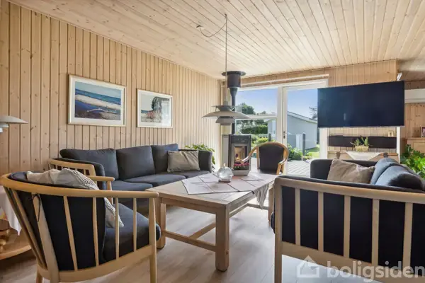 Stue med mørke sofasæt rundt om et træbord, pejs i midten, store vinduer med udsigt til en terrasse og have. Væggene har træpaneler, og billeder hænger dekorativt.